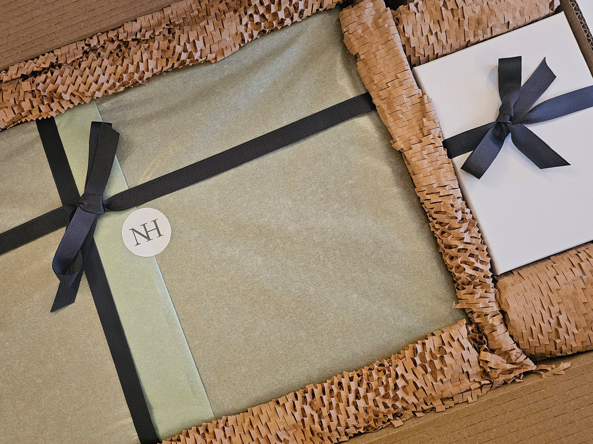 A chopping board gift wrapped in teal tissue paper and tied with a black ribbon and neat bow. Next to the gift wrapped board is a white gift box tied with a black ribbon. Both items are placed in cardboard packaging.