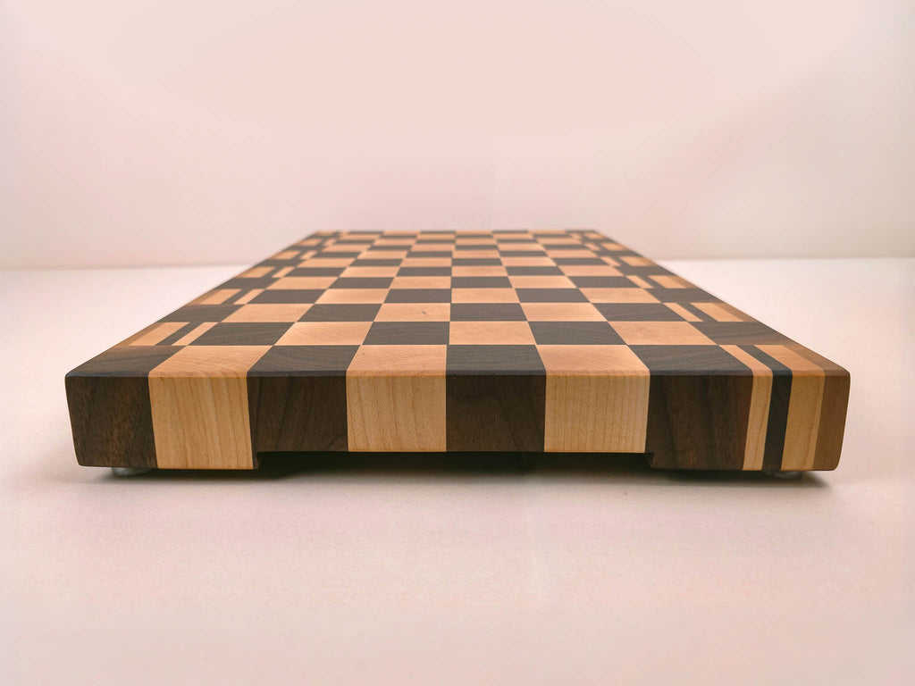 A rectangular wooden cutting board with a classic walnut and maple checkerboard pattern. This photo is showing the side view of the board to display it's underneath finger grooves from the side.