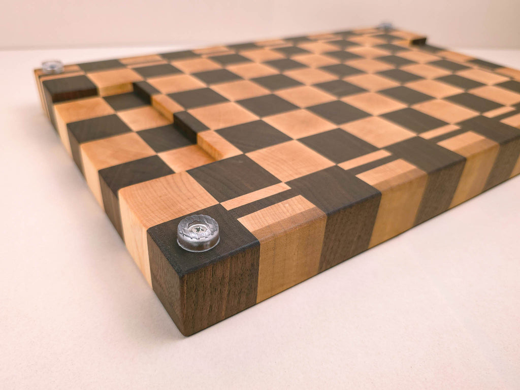 A rectangular wooden chopping board with a walnut and maple wood checkerboard pattern. This board is placed upside down on the kitchen counter to show it's underside finger groves and silicone feet.