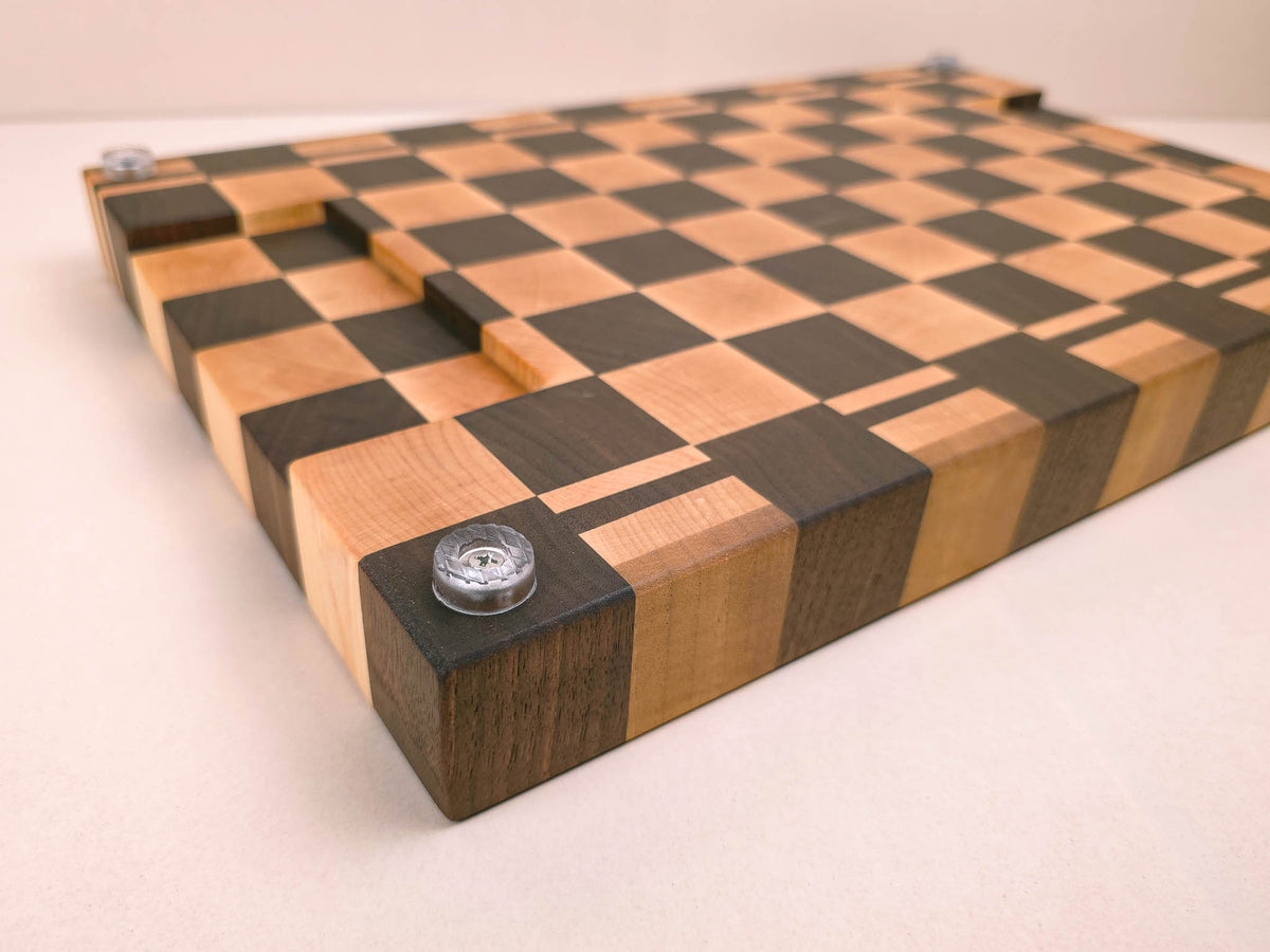 A rectangular wooden chopping board with a walnut and maple wood checkerboard pattern. This board is placed upside down on the kitchen counter to show it's underside finger groves and silicone feet.