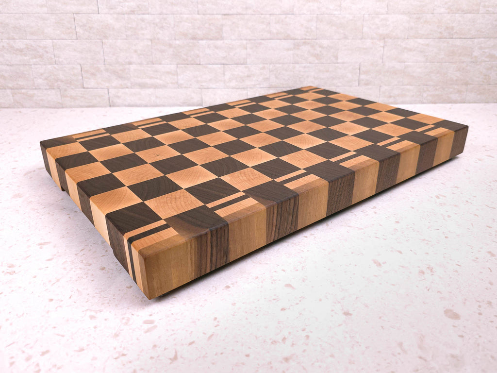 A large rectangular wooden cutting block with a checkerboard pattern in maple and dark walnut wood, shown on a light kitchen countertop against a light brick wall.