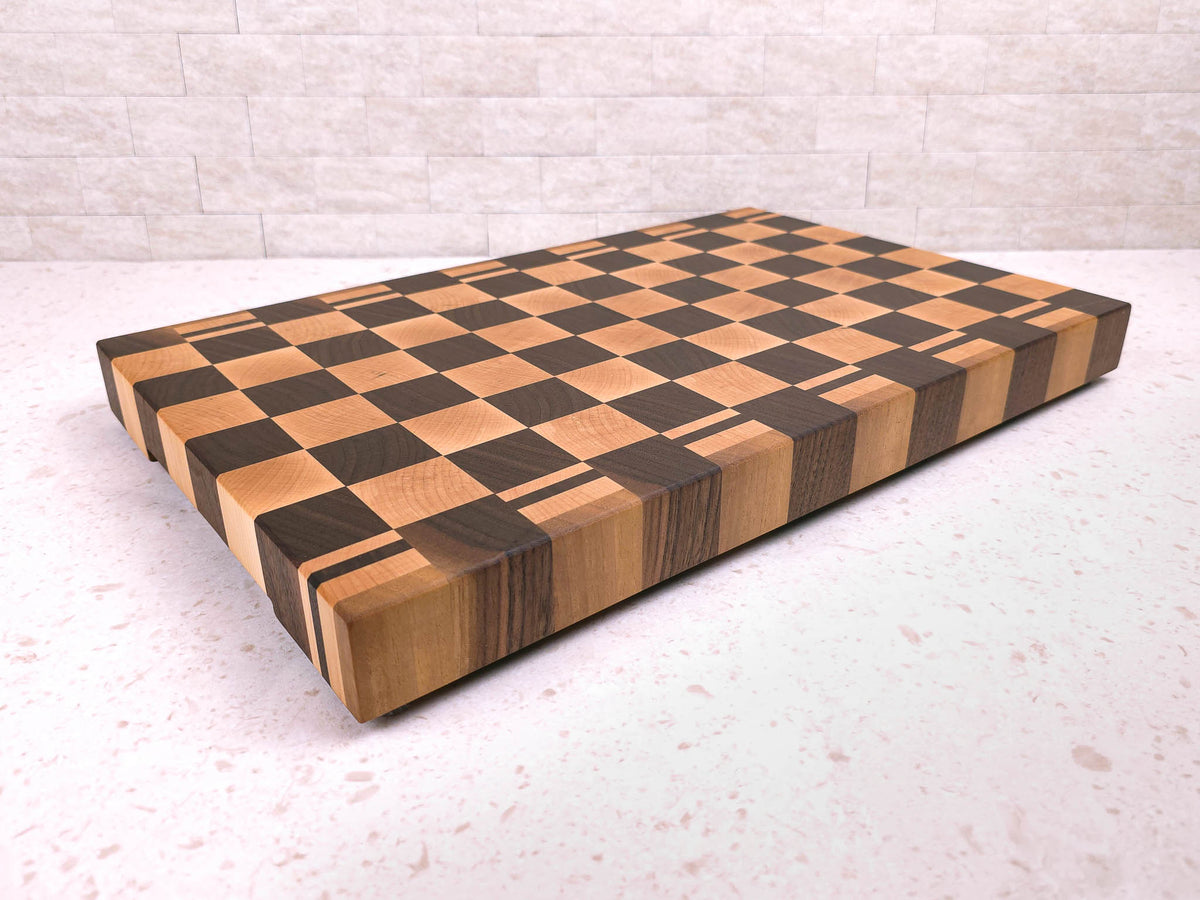 A large rectangular wooden cutting block with a checkerboard pattern in maple and dark walnut wood, shown on a light kitchen countertop against a light brick wall.