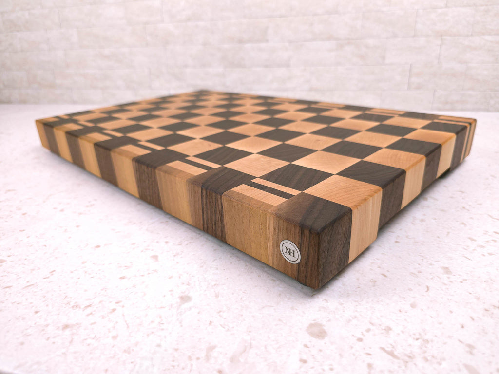 A wooden end-grain cutting block with a checkerboard pattern made of alternating maple and dark walnut wood blocks, photographed on a light stone surface against a white kitchen tiled backdrop.
