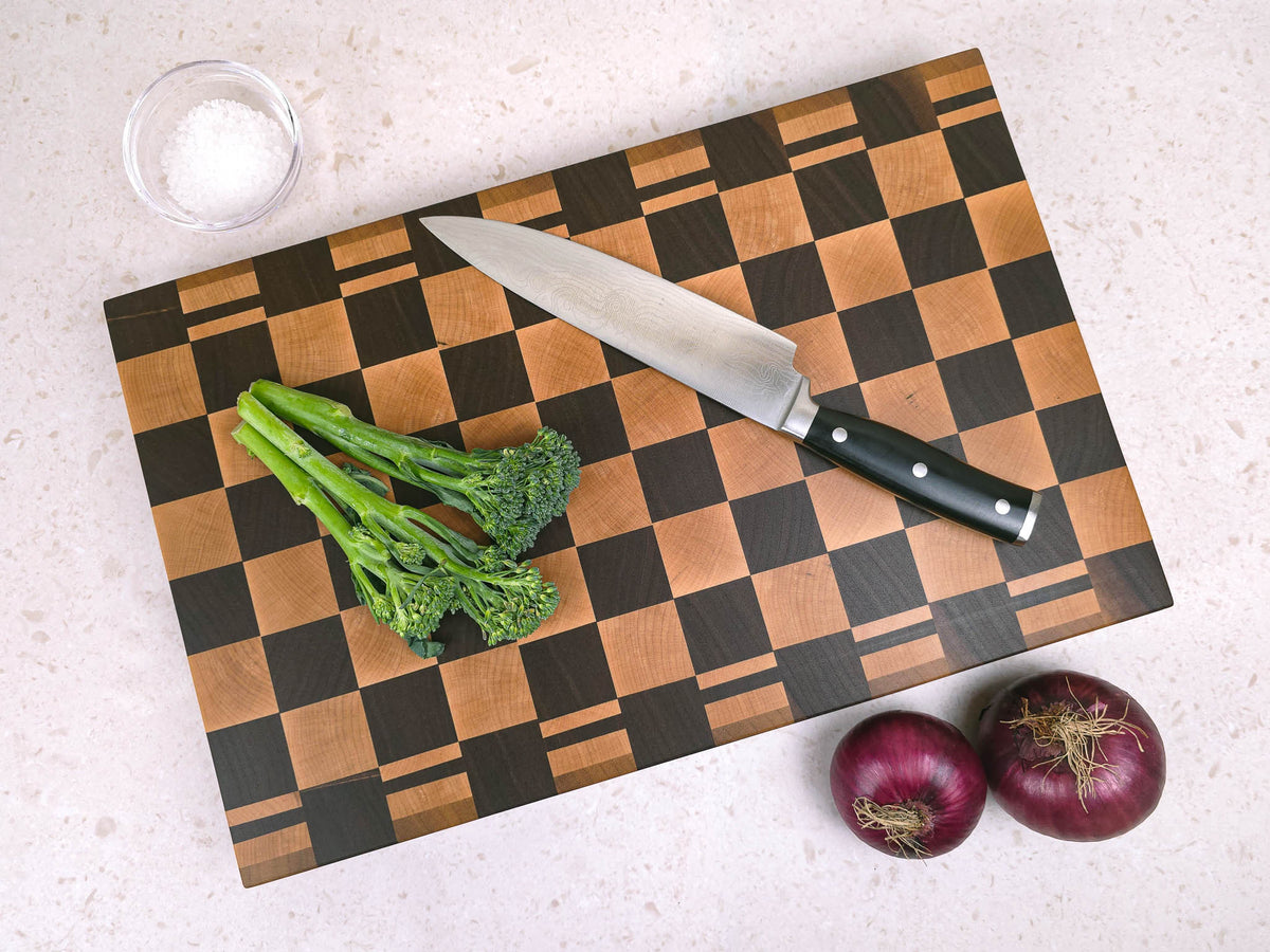 A large wooden chopping board with a walnut and maple wood checkerboard pattern, styled with a chef's knife, broccoli, and red onions on a light beige countertop.