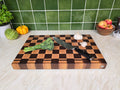A rectangular wooden cutting board with a dark and light checkerboard pattern sits on a light marble countertop. A chef's knife with a black handle rests on the board, alongside a stalk of broccoli, a garlic bulb, and a small white bulb.