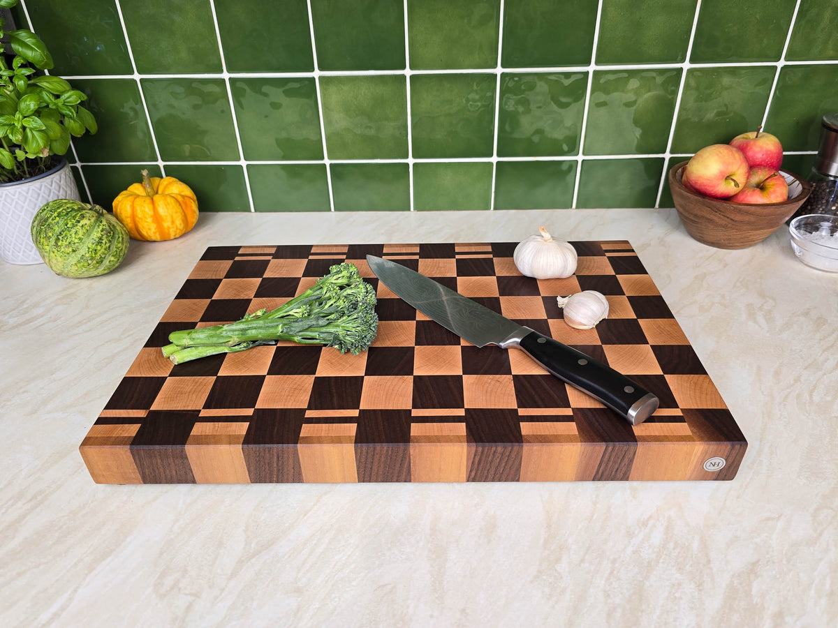 A rectangular wooden cutting board with a dark and light checkerboard pattern sits on a light marble countertop. A chef's knife with a black handle rests on the board, alongside a stalk of broccoli, a garlic bulb, and a small white bulb.
