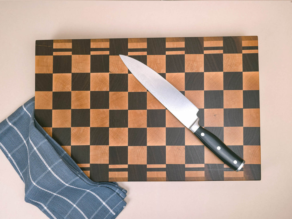 A chef's knife with a black handle rests diagonally on a luxury end-grain wooden cutting block with a maple and walnut checkerboard pattern. Set against a solid beige background with a navy tea towel placed over one corner of the chopping board.