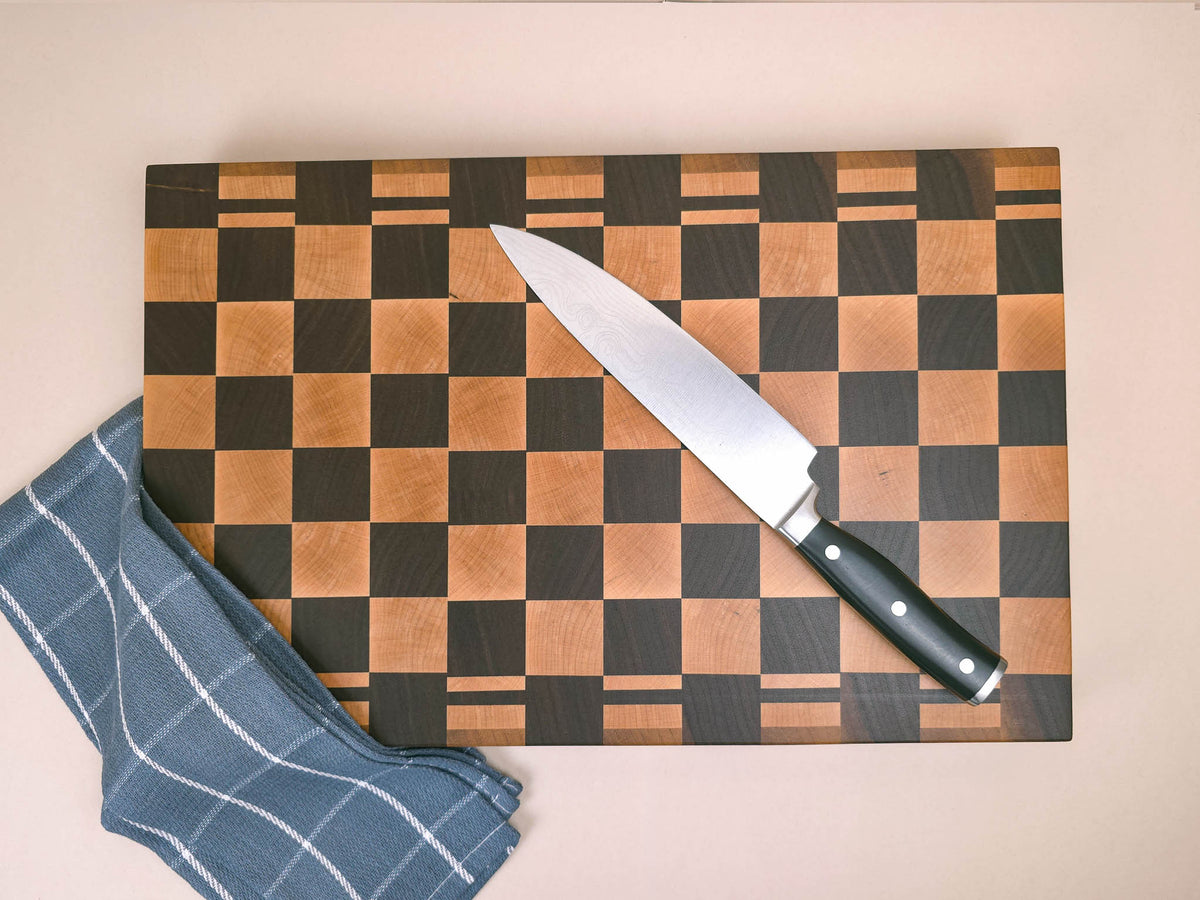 A chef's knife with a black handle rests diagonally on a luxury end-grain wooden cutting block with a maple and walnut checkerboard pattern. Set against a solid beige background with a navy tea towel placed over one corner of the chopping board.